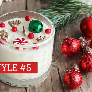 May include: A clear glass candle with a textured honeycomb pattern filled with white wax, topped with miniature gingerbread figures, a red and white peppermint candy, red spheres, and a green ornament. A red rectangular label reads STYLE #5.