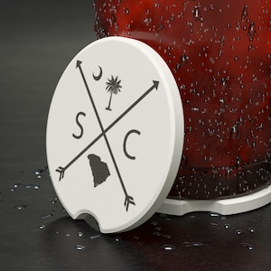 Puede incluir: Un posavasos redondo blanco con un diseño negro que presenta las letras "S" y "C", una luna creciente, una palmera y un contorno estatal. Una copa de líquido rojo está al lado del posavasos.