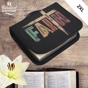 May include: Black Bible cover with the word "LOVE" in a cross design, featuring Bible verses. The cover has a zip closure and is labelled "2XL". A white flower and an open Bible are in the foreground.