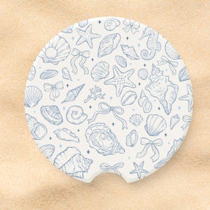 May include: A round, white car coaster featuring a blue seashell pattern. The design includes various seashells, starfish, bows, and a seahorse. The coaster has a small notch for easy removal from the car's cup holder.