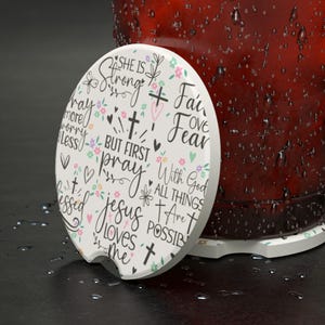 May include: White car coaster with a floral and script design. The coaster features the phrases "She is Strong", "But First Pray", and "Jesus Loves Me" in black script, with crosses and hearts. The coaster is next to a red glass.