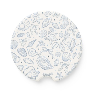 May include: A round, off-white car coaster featuring a blue line art pattern of seashells, starfish, bows, and a seahorse. The coaster has a small notch for easy removal from a car cup holder.