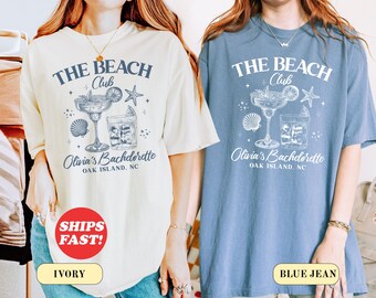 May include: Ivory and blue jean-colored t-shirts with the text "THE BEACH Club" and "Olivia's Bachelorette, OAK ISLAND, NC" printed on the front. The design includes a cocktail, seashell, and starfish graphic. The ivory shirt has a red "SHIPS FAST!" sticker.