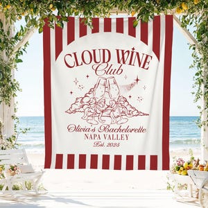 May include: A white banner with red stripes and text that reads "Cloud Wine Club" with a wine glass illustration. The banner also says "Olivia's Bachelorette, Napa Valley, Est. 2025". The banner is hanging on a white wooden structure with a beach background.