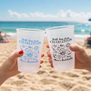 May include: Two translucent plastic cups featuring the text With love from CHARLESTON and Olivia's Bachelorette. One cup has blue ink graphics of a seashell, starfish, sunglasses, and a drink, while the other cup features the same graphics in black ink.