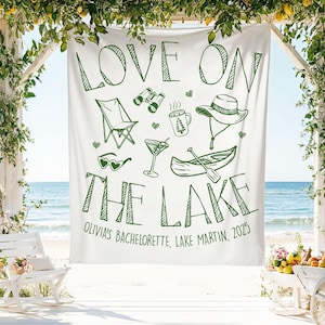 May include: White banner with green text and illustrations, reading "LOVE ON THE LAKE" with images of a chair, binoculars, coffee mug, hat, sunglasses, martini, and canoe. The banner also includes "Olivia's Bachelorette, Lake Martin, 2025".