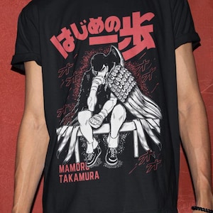May include: Black t-shirt with a red and white manga-style graphic. The graphic features a seated figure with a wing-like design and Japanese text. The text "MAMORU TAKAMURA" is printed below the illustration.