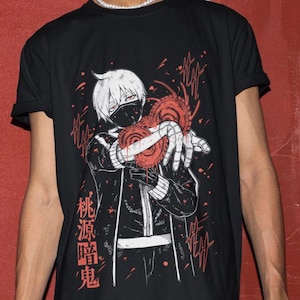 Puede incluir: Camiseta negra con una ilustración de estilo manga de un personaje con pelo blanco y una máscara, sosteniendo dos orbes de energía rojos. Texto japonés impreso en el lado izquierdo del gráfico. La camiseta tiene mangas cortas.