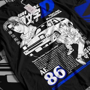 Puede incluir: Camiseta negra con una ilustración de estilo manga de un coche y dos figuras. El diseño incluye texto japonés y el número 86 en blanco y azul. La camiseta también tiene texto que describe las características del coche.