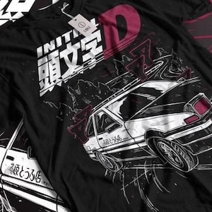 Peut inclure: T-shirt noir avec un motif d'une voiture blanche et noire dérivant sur une route de montagne. Le design comprend du texte japonais et la lettre "D" en bordeaux. Une étiquette indique la taille L.