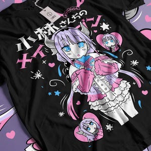 Puede incluir: Camiseta negra con una colorida ilustración de estilo anime. El diseño presenta un personaje con ojos azules, cuernos y pelo largo, rodeado de corazones, estrellas y texto japonés. El personaje lleva un atuendo rosa y azul.
