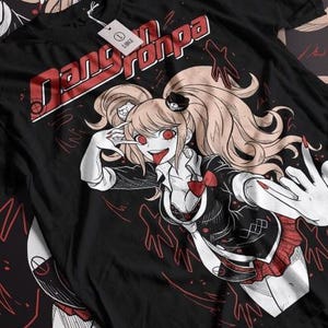 Op de afbeelding: Zwart t-shirt met de tekst "Danganronpa" in rood en wit. Het shirt toont een gedetailleerde anime-illustratie van een personage met blonde staartjes, rode ogen en een zwart-rode outfit. Er is een label aan bevestigd.