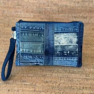 May include: A denim clutch with a wrist strap. The clutch is made from patches of blue jean material in various shades of blue. A dark blue zipper and wrist strap complete the design. The clutch is rectangular in shape.