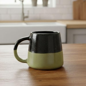 May include: A two-toned ceramic mug featuring a dark charcoal black upper section and a muted olive green lower section. The mug has a wide, rounded handle on the left side and a smooth, slightly tapered cylindrical body with a speckled texture.