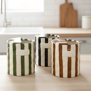May include: Three ceramic containers with vertical stripes in green and white, black and white, and brown and white. Each has a rectangular accent and a curved top. The containers are cylindrical and sit on a light-colored surface.