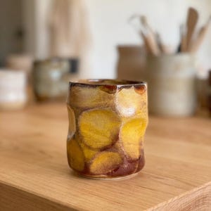 May include: A cylindrical ceramic cup featuring a hand-thrown, dimpled texture. The surface is finished with a mottled glaze in shades of deep amber, golden yellow, and dark brown, creating a layered, organic appearance with a glossy, reflective sheen.