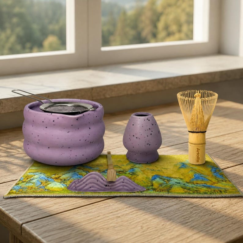 May include: A matcha tea set featuring a lavender ceramic bowl with a stainless steel strainer, a matching ceramic tea bowl, a bamboo whisk, and a decorative mat with a mountain design. The set is arranged on a wooden surface.
