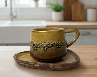 May include: A ceramic mug and matching saucer set featuring a two-tone mustard yellow and dark brown glaze. The mug has a textured, bumpy band around its center and a curved handle. The saucer is irregularly shaped with a smooth, flat surface.