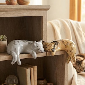 May include: Two ceramic cat figurines resting on a wooden shelf edge. One cat is light gray with a distressed, speckled finish, while the other is a warm tan color with dark brown mottled spots. Both figures are sculpted in a reclining pose.