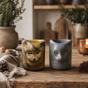 May include: Two ceramic mugs featuring sculpted cat faces on the front. One mug is a mustard yellow with dark brown markings, while the other is a solid glossy grey. Both mugs have a cylindrical shape with a molded feline ear detail at the rim.