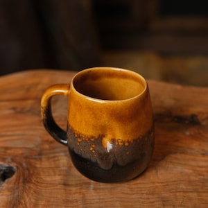 May include: A ceramic mug featuring a two-tone glaze with a warm golden-yellow upper section that transitions into a deep, dark brown base. The mug has a rounded body, a smooth rim, and a curved handle on the side, showcasing a reactive finish.