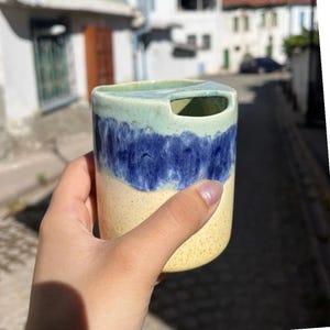 May include: A cylindrical ceramic cup featuring a speckled cream base, a wide band of deep blue glaze, and a light green rim. The top edge of the cup includes a distinct cutout notch, creating a unique opening on one side of the vessel's rim.