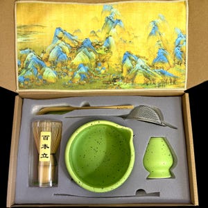 May include: A matcha tea set in a box, including a green ceramic bowl, whisk, scoop, sifter, and a whisk holder. The set has a yellow and blue landscape design on the lid. The whisk is in a clear container with Japanese text.