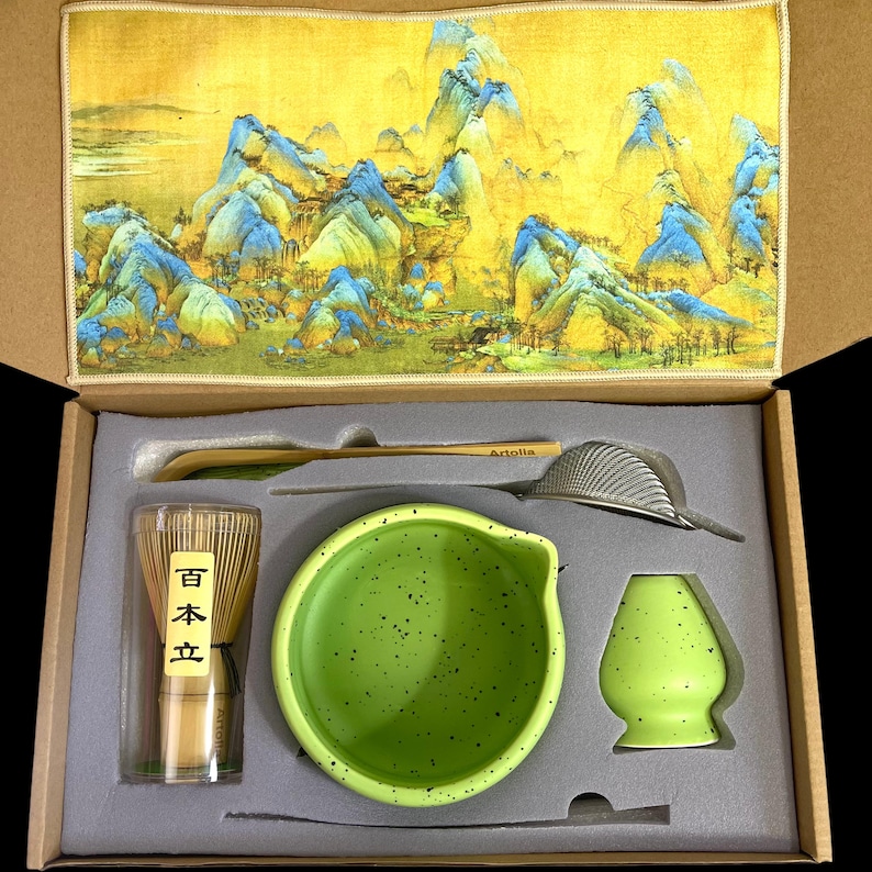 May include: A matcha tea set in a box, including a green ceramic bowl, whisk, scoop, sifter, and a whisk holder. The set has a yellow and blue landscape design on the lid. The whisk is in a clear container with Japanese text.