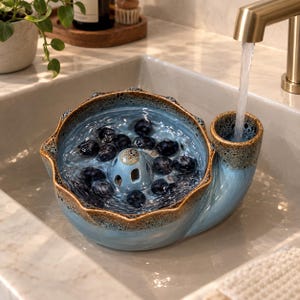 May include: A light blue ceramic berry colander with a scalloped rim and a brown speckled glaze. The bowl features a central drainage cone and a side handle, currently filled with fresh dark blueberries being rinsed under a gold faucet.