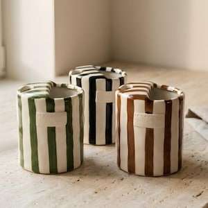 May include: Three cylindrical ceramic holders with vertical stripes in forest green, black, and brown. Each holder features a white rectangular embossed label on the front and a notched rim design, providing a structured look for storage.