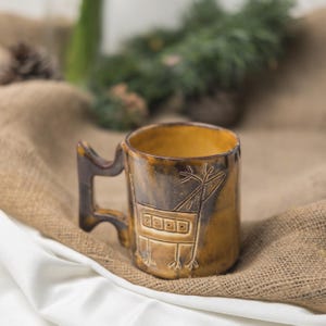 May include: A cylindrical ceramic mug with a glossy mustard yellow and dark brown glaze. The exterior features a hand-etched line drawing of a stylized four-legged animal with antlers. The mug has a unique angular handle with a cutout design.