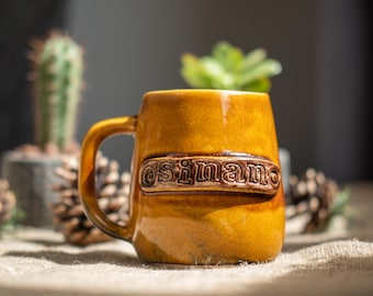 May include: A mustard yellow ceramic mug with a glossy finish and a single handle. A raised rectangular plaque on the front features the embossed text "sinan" in lowercase letters. The mug has a slightly tapered shape and a smooth, glazed surface.