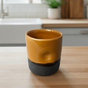 May include: A cylindrical ceramic mug featuring a two-tone design with a glossy mustard yellow upper section and a matte charcoal black base. The exterior surface includes a single thumb-sized indentation near the top rim for a secure grip.