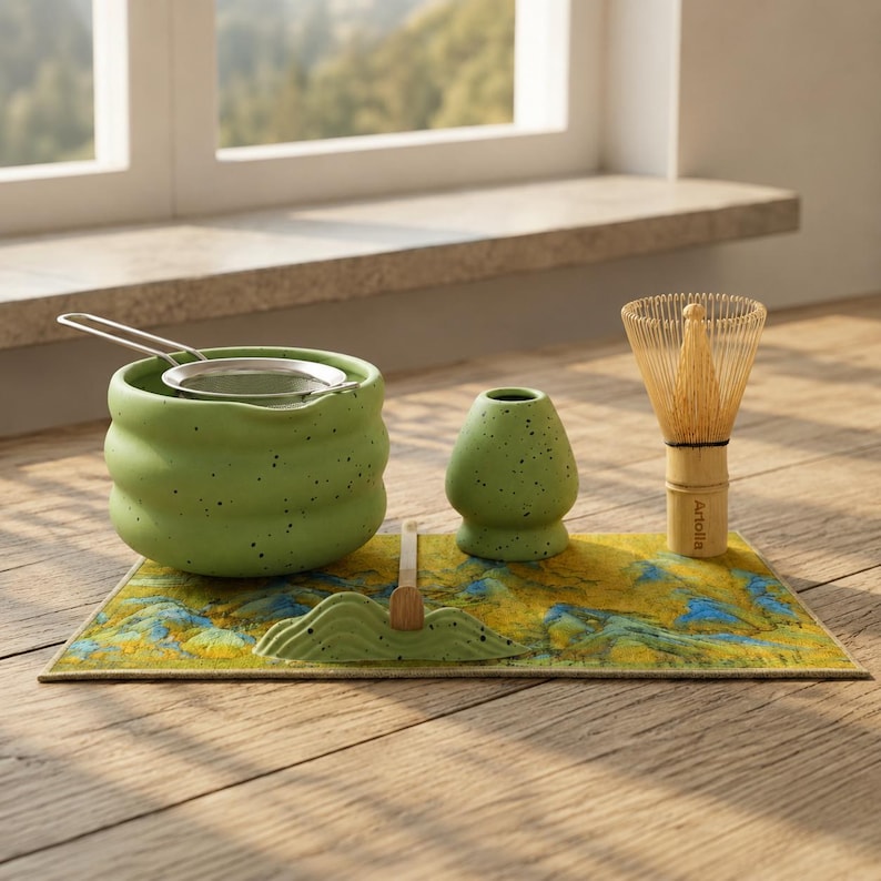 May include: A matcha tea set featuring a green ceramic bowl with a stainless steel strainer, a whisk, a whisk holder, and a small ceramic container. The set is on a decorative mat with a yellow and blue landscape design. The whisk has a bamboo handle.