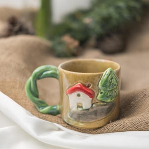 May include: A tan ceramic mug featuring a sculpted white house with a red roof and a green tree on the exterior. The mug has a unique twisted green handle and a glossy finish, showcasing a three-dimensional design that wraps around the side.
