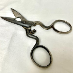 May include: A pair of vintage-style metal scissors with a dark, weathered finish. The tool features two oval finger loops, a central screw pivot, and a small brass-colored adjustment knob located between the handle and the pointed blades.