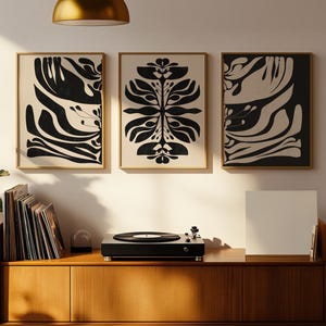 Może przedstawiać: Trzy oprawione abstrakcyjne wydruki artystyczne z czarno-kremowymi wzorami wiszą nad drewnianą konsolą gramofonową. Czarny gramofon stoi na górze, ze stosem płyt winylowych po lewej stronie. Roślina doniczkowa znajduje się po lewej stronie.