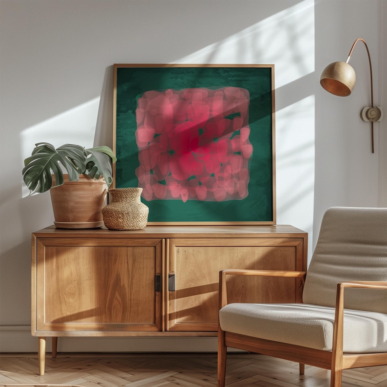 May include: A framed abstract art print with a dark green background and a central, textured, red design. The artwork is displayed above a wooden cabinet, with a potted plant and a woven vase. A modern chair and a wall-mounted lamp are also visible.