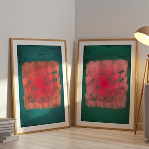 May include: Two framed abstract art prints. Each features a red and orange central design on a dark green background. The frames are light wood. The prints are leaning against a white wall.