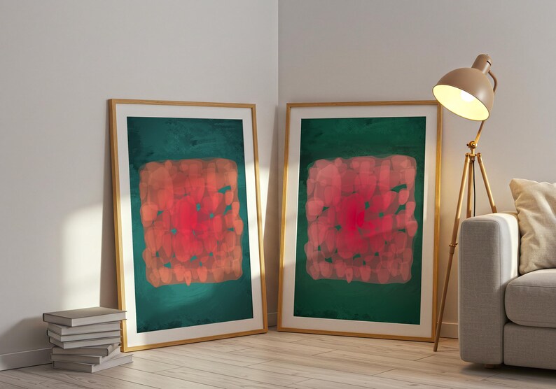 May include: Two framed abstract art prints. Each features a red and orange central design on a dark green background. The frames are light wood. The prints are leaning against a white wall.