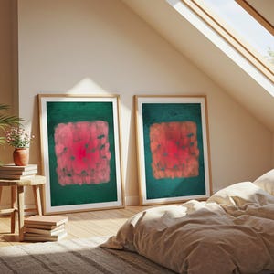 May include: Two framed abstract art prints. Each features a central, textured, red and pink square on a dark green background. The frames are light wood, and the art is displayed in a room with a bed and a skylight.