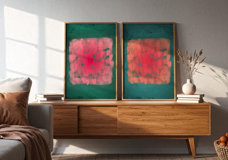 May include: Two framed abstract art prints. Each features a red and pink square design on a teal green background. The artwork is displayed on a wooden cabinet with a vase and books.