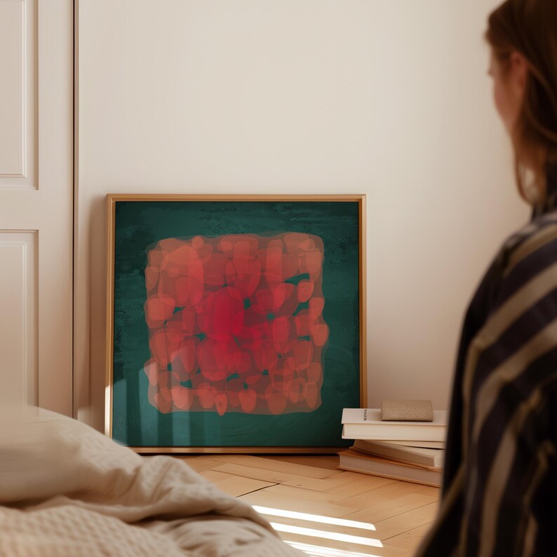 May include: Abstract art print in a gold frame. The artwork features a square composition of red and orange brushstrokes on a dark green background. The painting is displayed on a wooden floor, next to a stack of books.