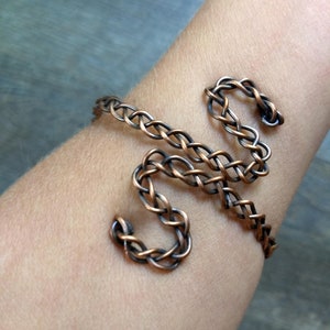 May include: A copper wire bracelet with a braided design. The bracelet is shaped like the letter "S" and is worn on a person's wrist.