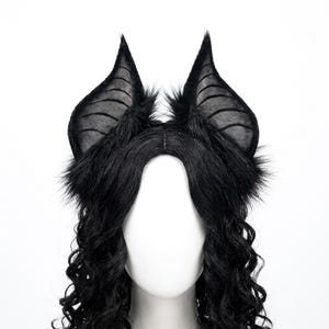 May include: A black headband featuring two large, pointed bat-like ears covered in faux fur. The ears have a thin, semi-transparent black fabric interior with visible stitched lines. The headband is worn on a white mannequin head with long, wavy black hair.