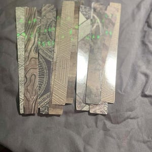 May include: A collection of rectangular bookmarks with various patterns and textures. The designs include wood grain, geometric shapes, and paisley. Some bookmarks have small, scattered green accents. The bookmarks are arranged on a gray surface.