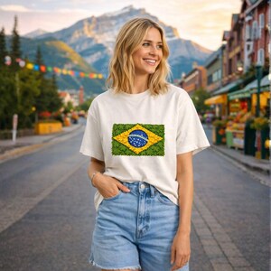 Può includere: Una maglietta bianca con un design floreale della bandiera brasiliana. La maglietta ha un girocollo e maniche corte. La persona indossa pantaloncini di jeans blu. Lo sfondo mostra una scena di strada con montagne.