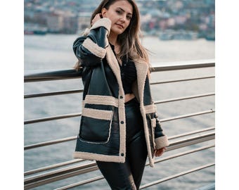 May include: A black faux leather jacket with cream-colored shearling trim and accents. The jacket features large pockets and a relaxed fit. The model is wearing black pants and a black top. The background shows a cityscape and water.