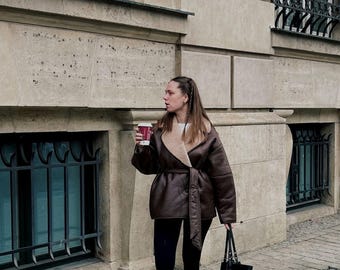 May include: A brown leather jacket with a cream-colored shearling lining and a matching belt. The jacket is worn over a white top and black leggings. A person is holding a red coffee cup and a black tote bag.