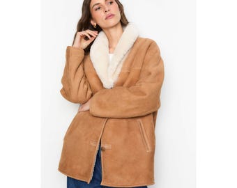 May include: A tan suede coat with a cream-colored shearling collar. The coat has a button closure and two front pockets. The model is wearing blue jeans. The coat is a classic outerwear piece.